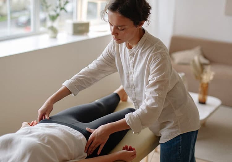 Massage therapist works on a client lying on a massage table in a bright, airy room, hands on the legs.